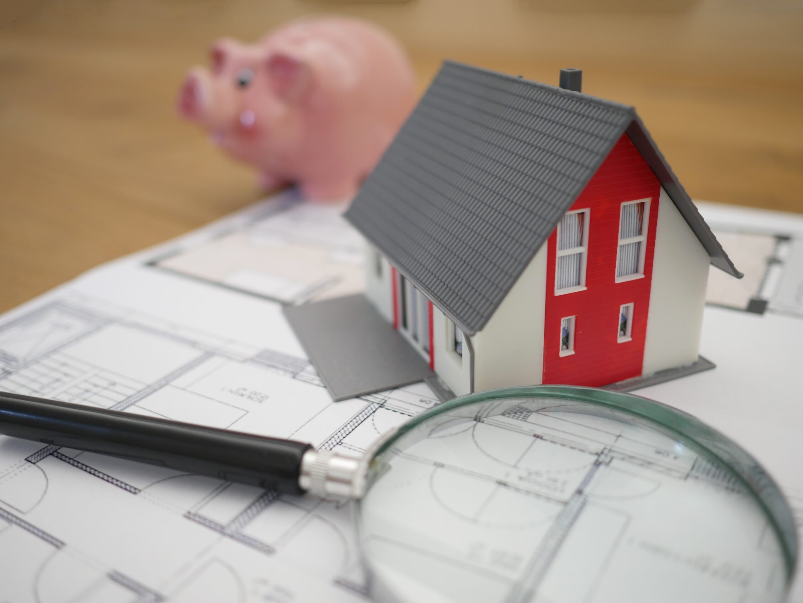 Imagen de una mini casa sobre sus planos, con una lupa y una hucha cerdo detrás.